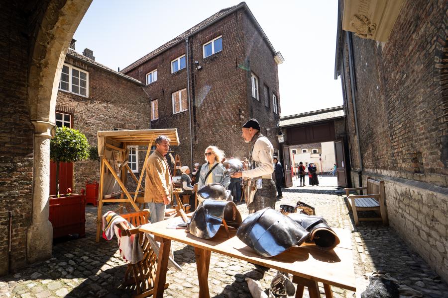 Mensen in middeleeuwse kostuums geven uitleg aan bezoekers over hetgeen op hun tafel ligt (helmen, eten ...)